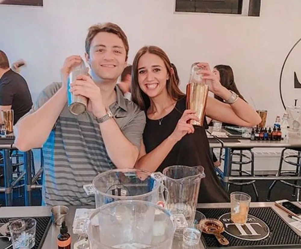 Two people are smiling at the camera holding cocktail shakers with various bar equipment and glasses on the table in front of them suggesting they are participating in a cocktail-making class or event