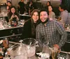 Two people are smiling at the camera holding cocktail shakers with various bar equipment and glasses on the table in front of them suggesting they are participating in a cocktail-making class or event