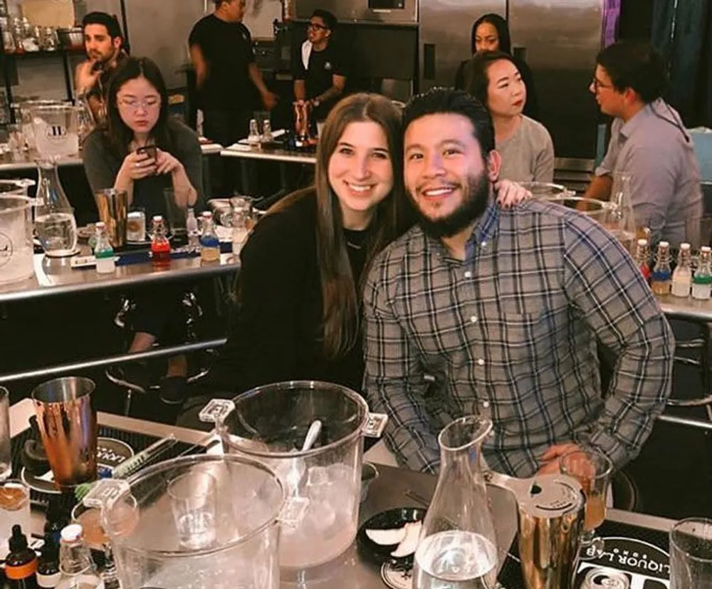 A smiling man and woman pose for a photo at a bar or mixology class surrounded by various glassware and bartending equipment