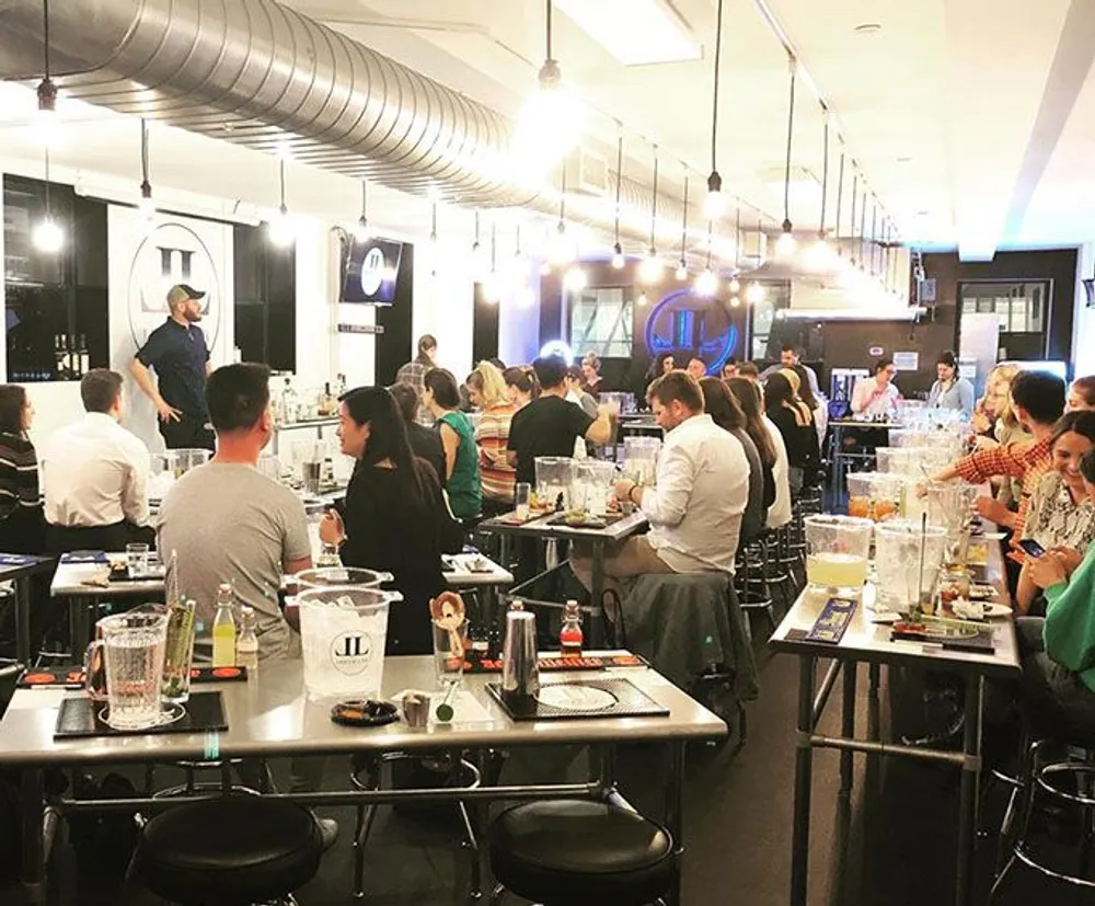 The image shows a lively cooking class with participants seated at individual cooking stations engaged in food preparation with an instructor at the front