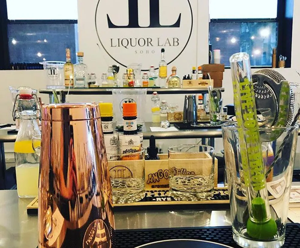 The image shows a well-organized bar setup with a variety of cocktail-making tools and ingredients on the counter in front of a backlit liquor shelf labeled LIQUOR LAB SOHO