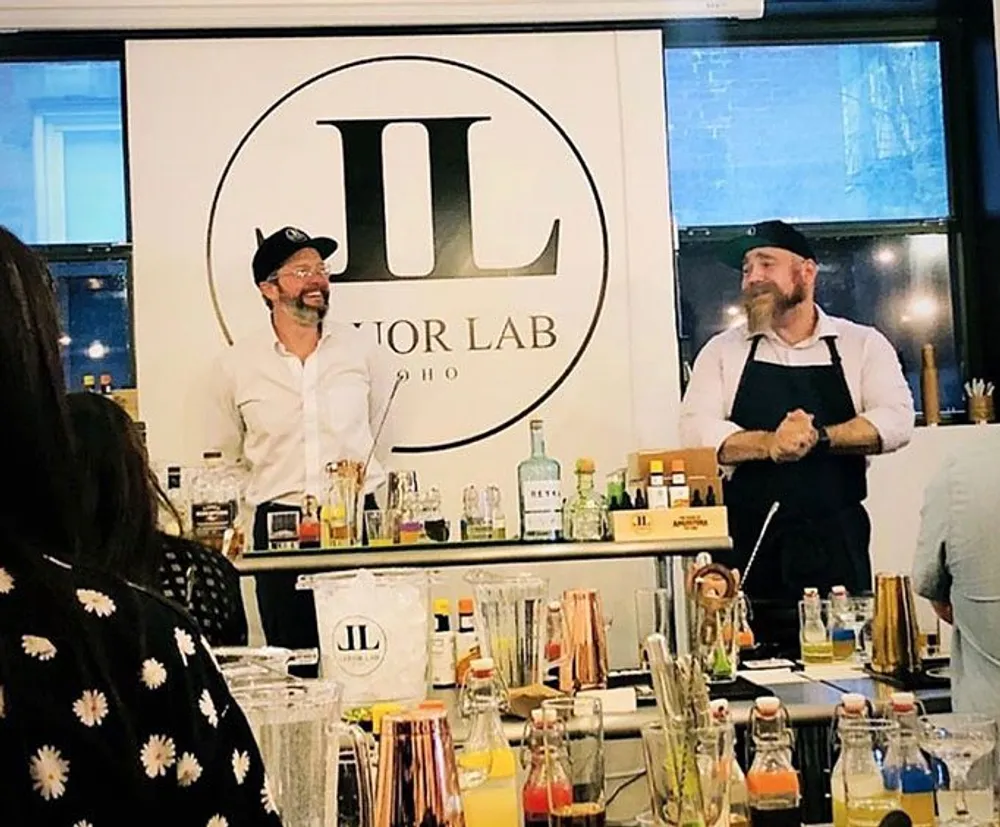 Two men are standing in front of a banner with the logo JL Creator Lab smiling and engaged in some sort of presentation or workshop involving various bottles and mixing equipment on a counter