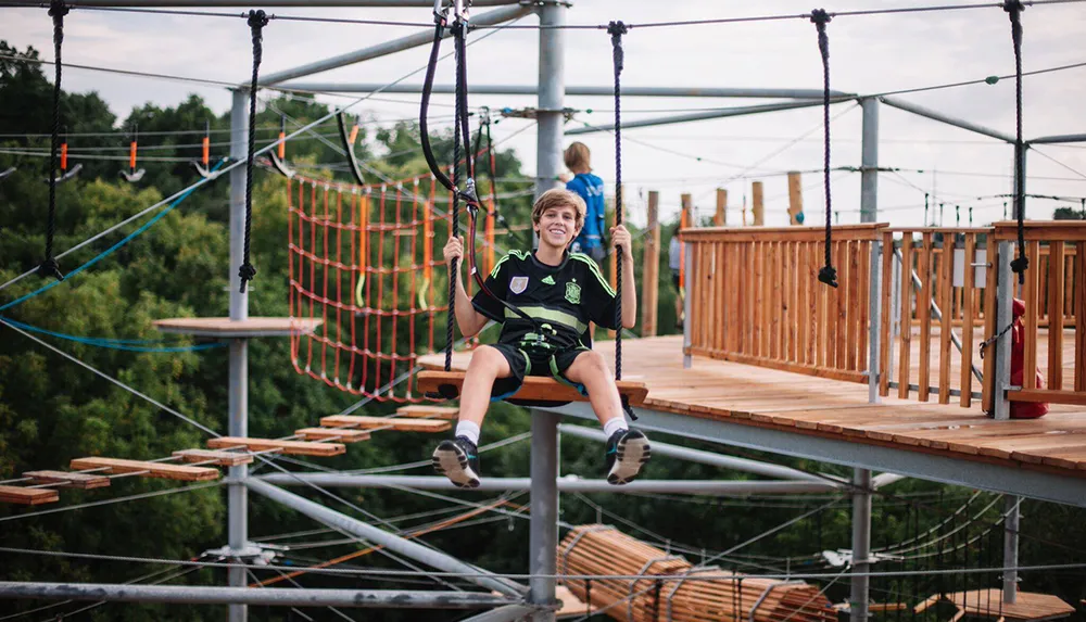 A smiling person is sitting on a swing at an aerial adventure park with obstacle courses in the background
