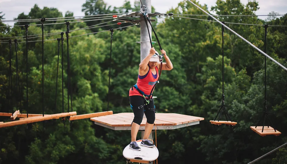 A person is engaged in an outdoor high ropes course navigating between platforms with the aid of a safety harness and wire-guided system above lush greenery