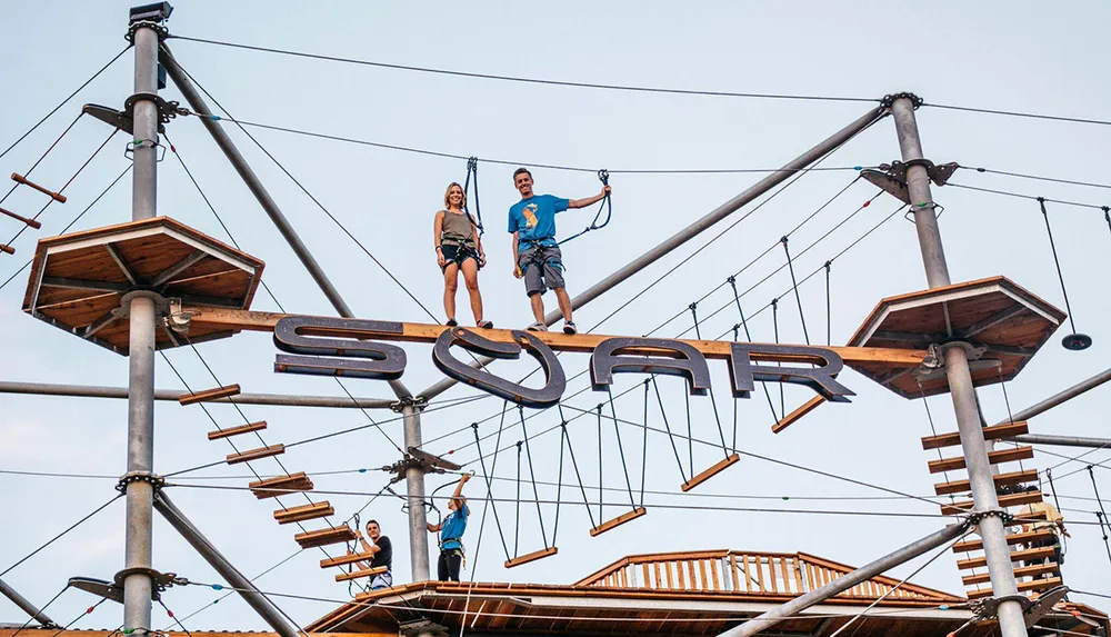 Two individuals are smiling on an elevated obstacle course with the word SOAR displayed prominently as they engage in an adventurous outdoor activity