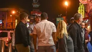 A group of people is gathered on a lively urban night street with neon signs and a sign for Korean Vets Bridge in the background.