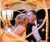 A couple in wedding attire shares an intimate kiss inside a brightly lit heart-shaped carriage structure at night