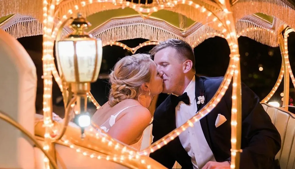 A couple in wedding attire shares an intimate kiss inside a brightly lit heart-shaped carriage structure at night