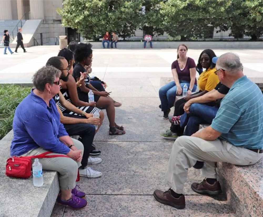 A diverse group of individuals is sitting on ledges in what appears to be a casual outdoor meeting or discussion