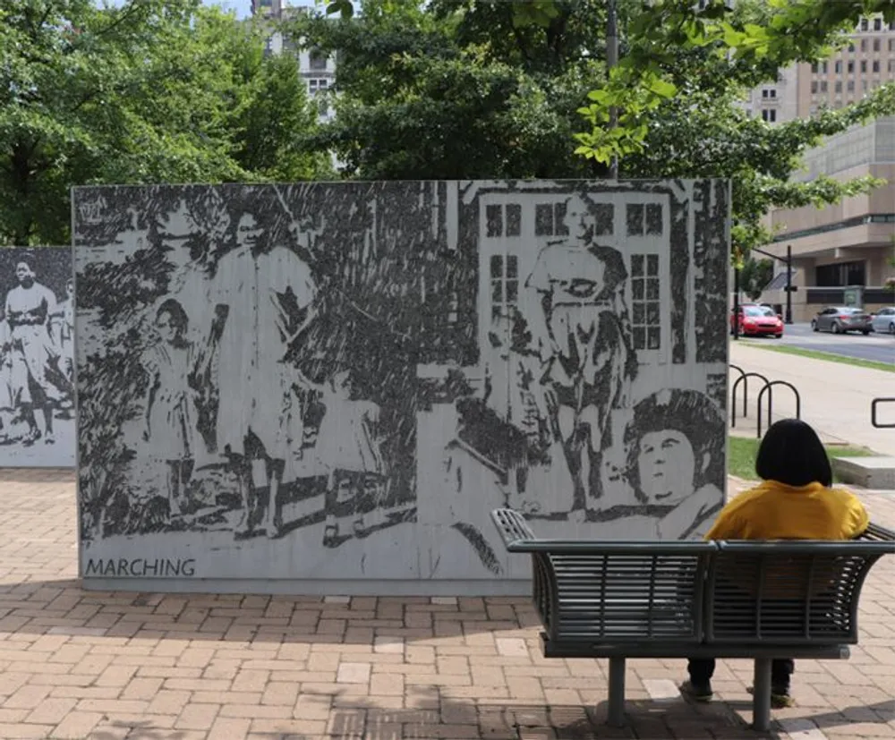 A person in a yellow top is sitting on a bench in front of a large black-and-white mural with the word MARCHING at the bottom left corner depicting historical images of individuals