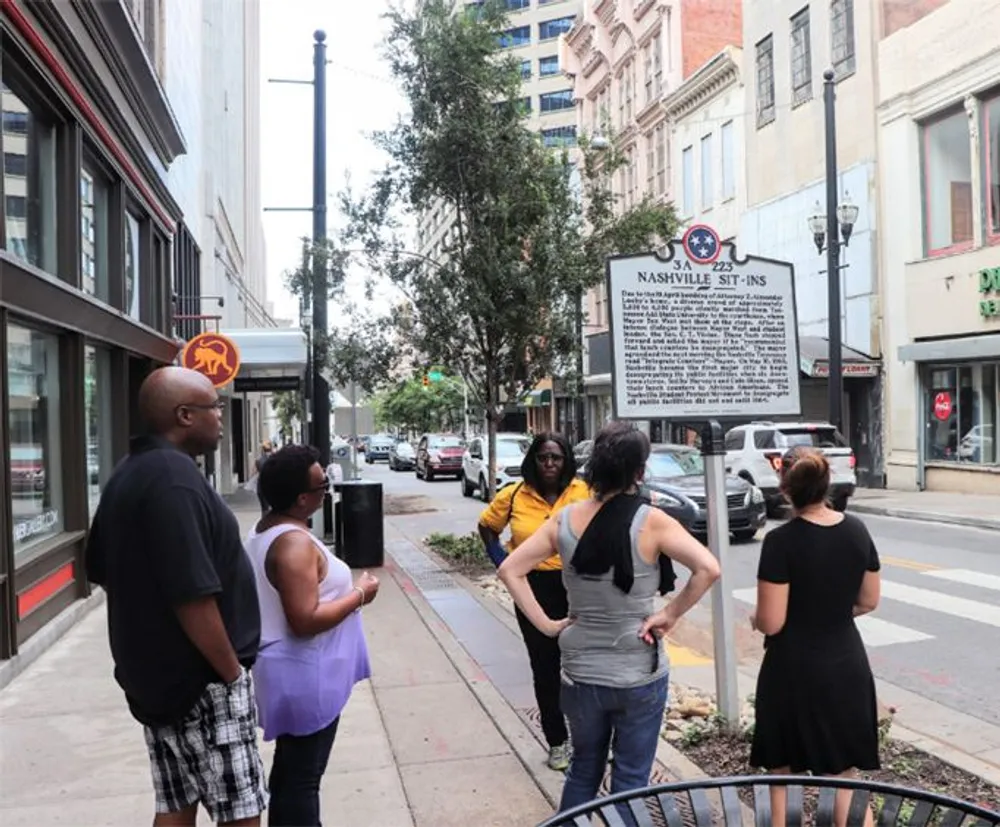 A group of people is standing on a sidewalk reading a historical marker titled Nashville Sit-Ins in an urban setting