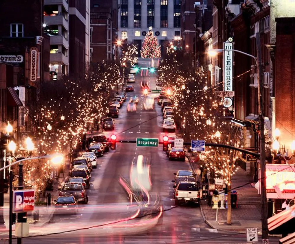 A bustling city street at dusk adorned with festive holiday lights and a large Christmas tree standing at the end captured with a long exposure to blur the moving vehicles