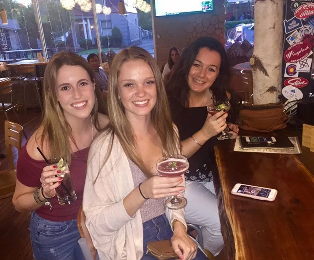 Three smiling women are posing for a photo while holding drinks in a bar-like setting