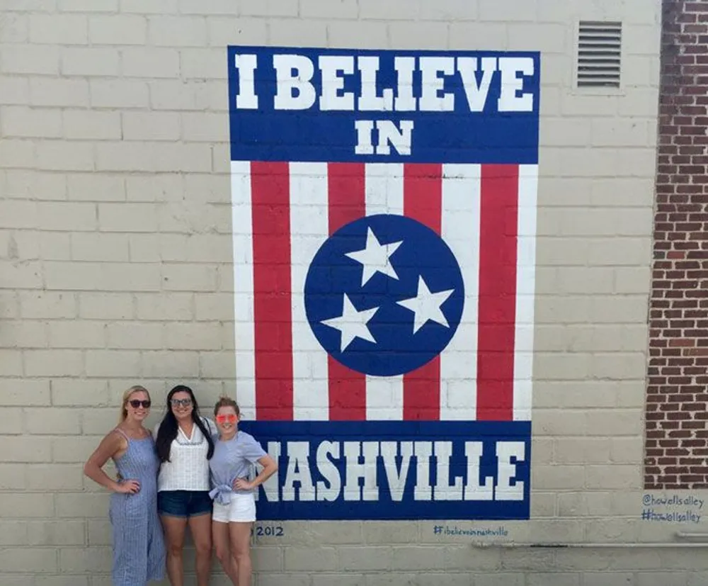 Three people are posing in front of a mural that states I BELIEVE IN NASHVILLE stylized with elements resembling the flag of Tennessee