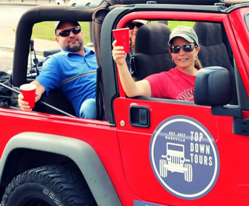 Two people are smiling and enjoying a ride in a red Jeep with a Top Down Tours Nashville decal on the side suggesting a leisurely tour experience