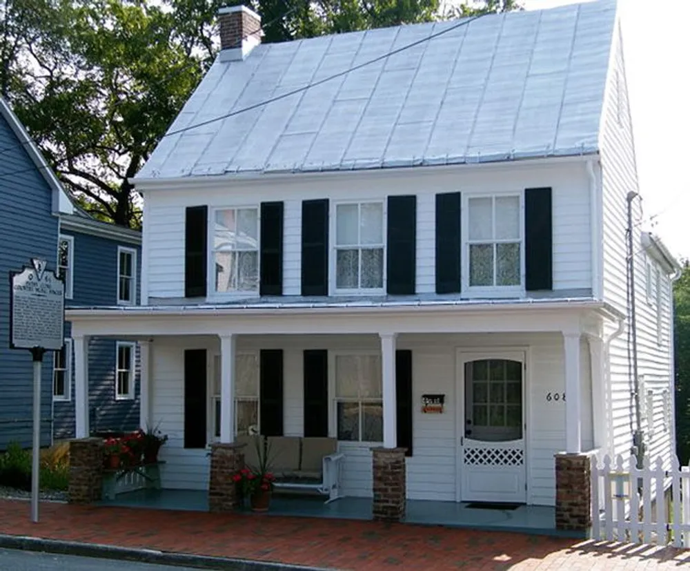 The image shows a quaint two-story white house with black shutters a metal roof and a historical marker next to it