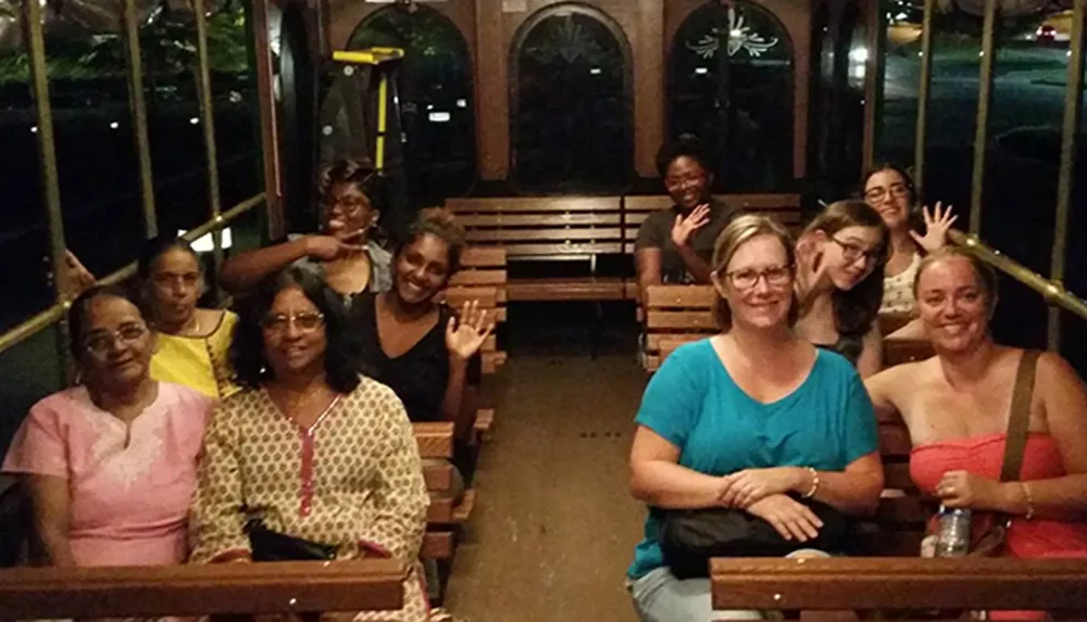 A group of smiling people is seated on a trolley or open-sided bus waving at the camera