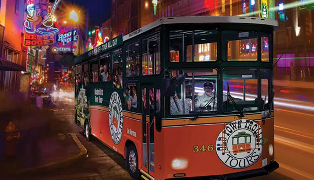 A tour bus designed to resemble a vintage trolley travels through a brightly-lit urban street at night filled with passengers