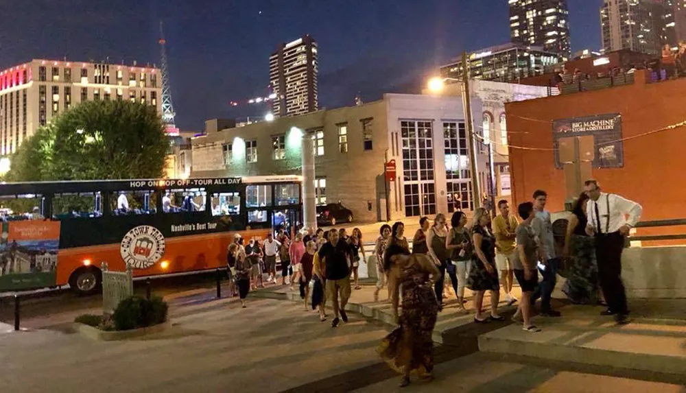 A group of people is walking on the sidewalk at dusk in an urban area with a hop-on hop-off tour bus parked nearby