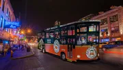A trolley bus full of passengers is on a city street at night with neon-lit establishments in the background.