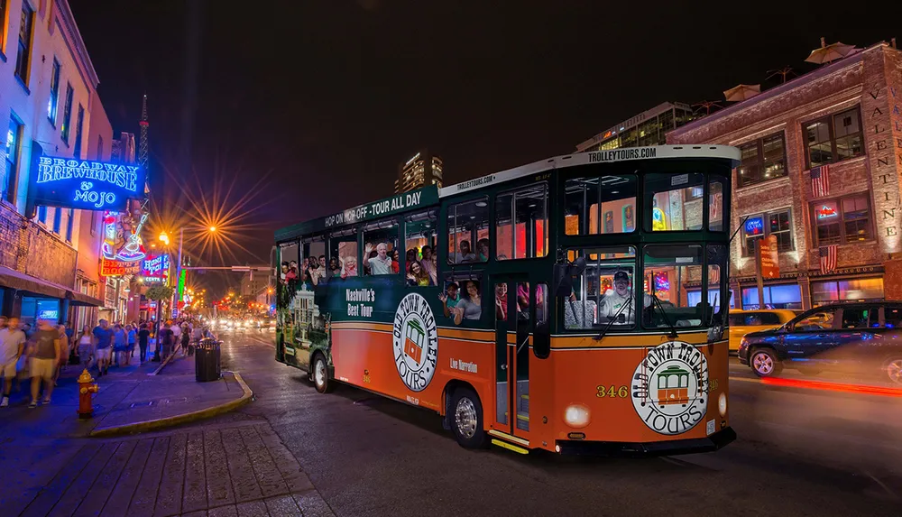 A trolley bus full of passengers is on a city street at night with neon-lit establishments in the background