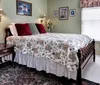 The image shows a neatly arranged bedroom with traditional furniture and decor featuring a floral bedspread vintage-style chairs and framed pictures on soft green walls