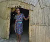 A smiling child is standing in the doorway of a thatch-roofed hut