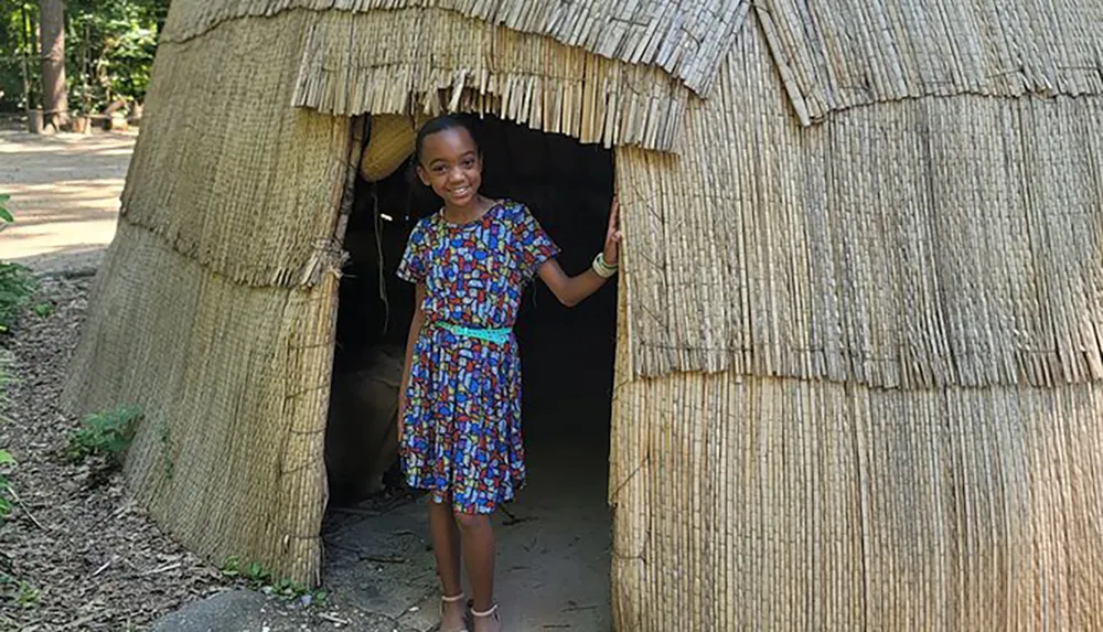 A smiling child is standing in the doorway of a thatch-roofed hut