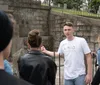 A person in a white t-shirt reading junket is standing near a metal gate and talking to others whose backs are turned to the camera