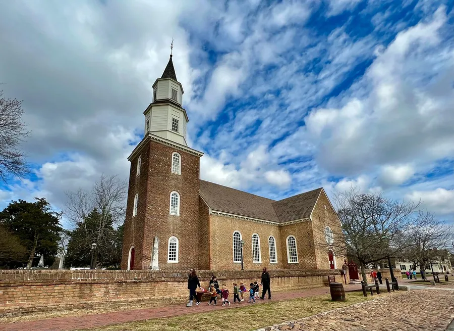 People visit the grounds of an historic brick church with a tall white steeple under a partly cloudy sky.