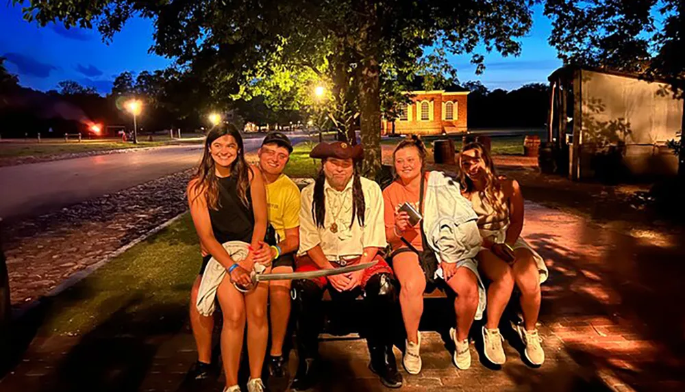 Four individuals are posing for a photo at night on a bench one dressed in historical period clothing with warm lighting and trees in the background