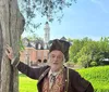 A person wearing historical clothing is posing next to a tree with a colonial-style building and tower in the background
