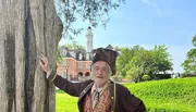 A person wearing historical clothing is posing next to a tree with a colonial-style building and tower in the background.