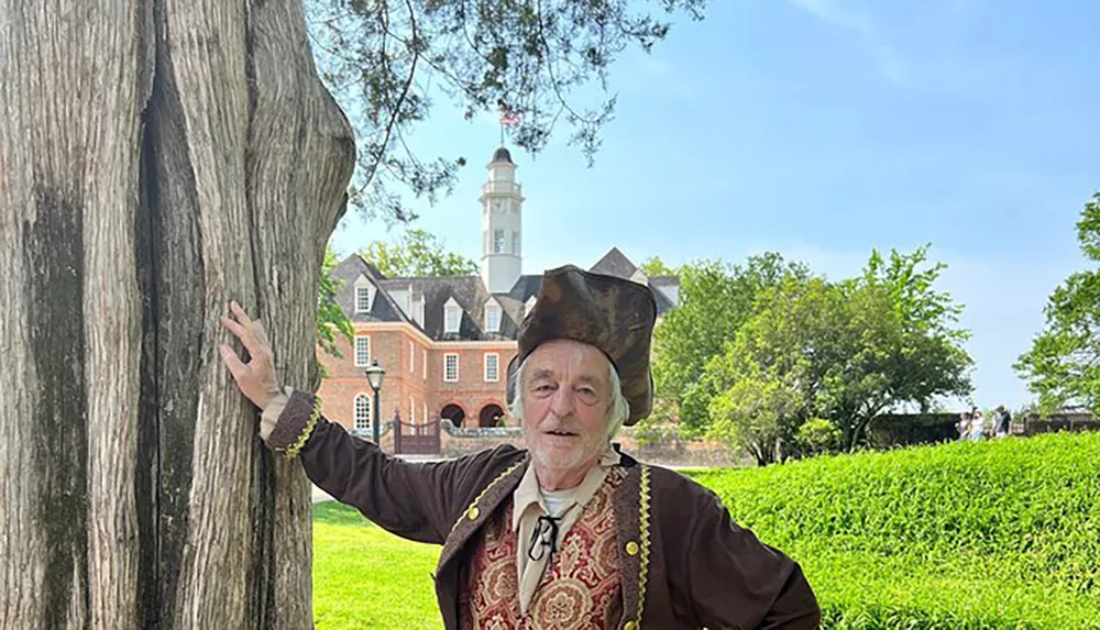 A person wearing historical clothing is posing next to a tree with a colonial-style building and tower in the background