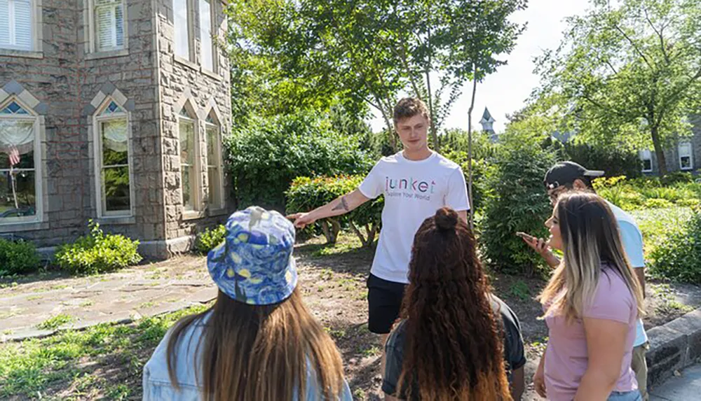 A person is standing and pointing something out to a group of attentive listeners outdoors near a stone building