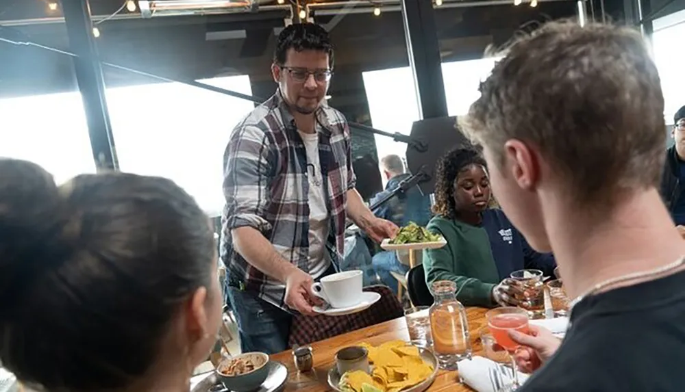 A person is serving food at a busy dining table where several other people are seated with drinks and plates in front of them