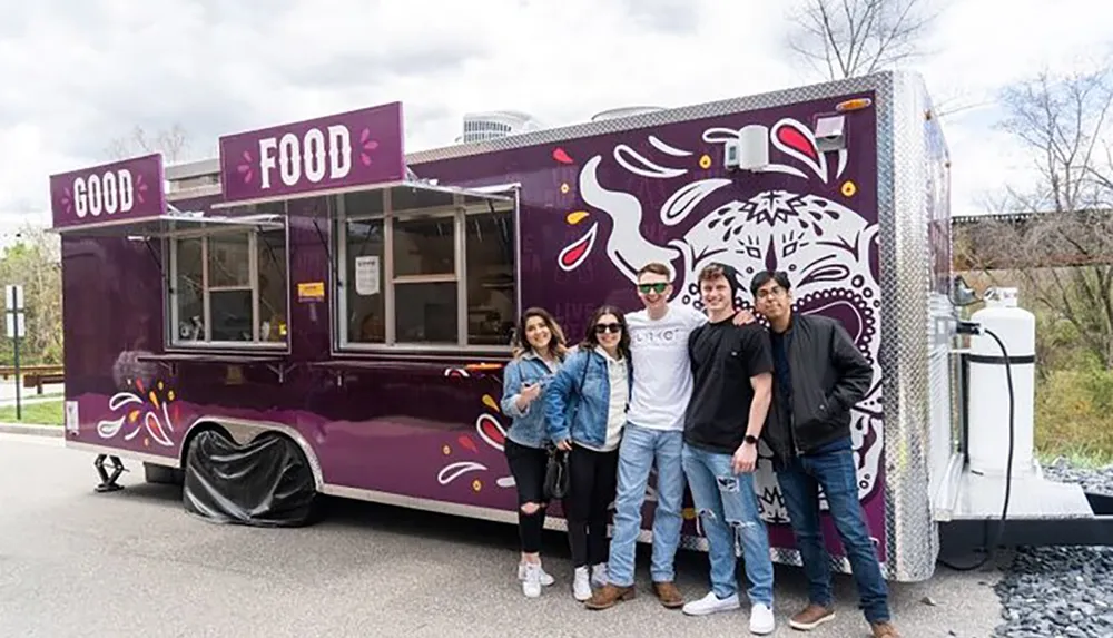 A group of five people are smiling and posing in front of a purple food truck with the words GOOD FOOD written on it