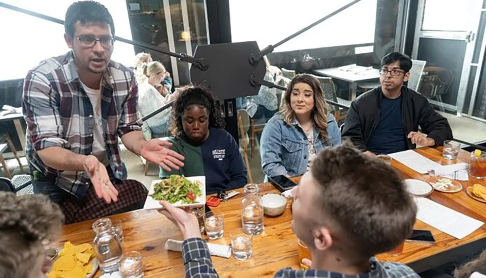 A group of people is sitting around a table in a casual dining setting with various expressions while a man standing gestures emphatically possibly engaged in storytelling or making a point