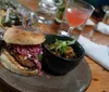 The image showcases a gourmet burger with a side of what appears to be a bean dish and a pink cocktail on a wooden restaurant table