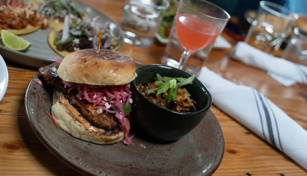 The image showcases a gourmet burger with a side of what appears to be a bean dish and a pink cocktail on a wooden restaurant table