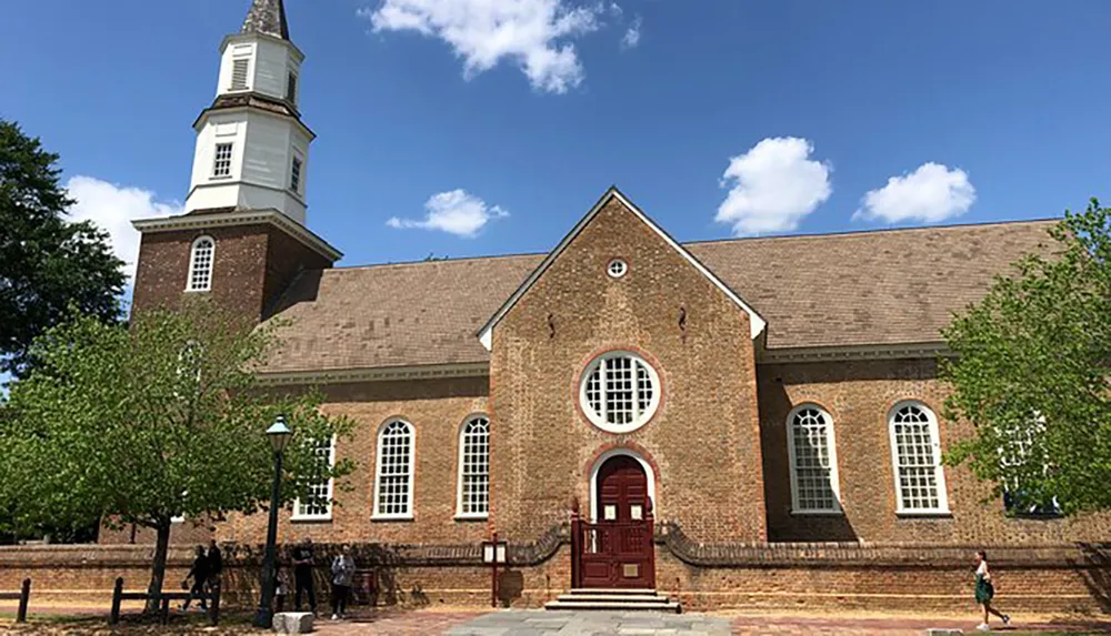 The image shows a traditional brick church with arched windows and a steeple against a blue sky with people walking by in the foreground