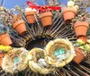 A man and a woman are smiling while standing beside a large ornate wreath adorned with various plants and decorations displayed outside a building with a brick step and railings