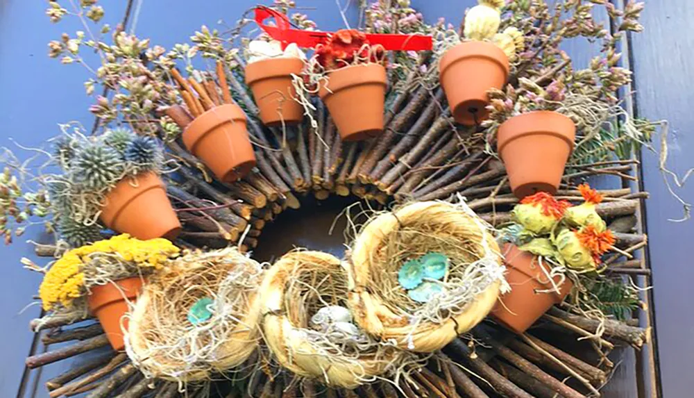 This image shows a decorative wreath made of twigs featuring small terracotta pots dried flowers and birds nests with eggs all arranged in a circular pattern against a blue door