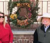 A man and a woman are smiling while standing beside a large ornate wreath adorned with various plants and decorations displayed outside a building with a brick step and railings