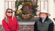 A man and a woman are smiling while standing beside a large, ornate wreath adorned with various plants and decorations, displayed outside a building with a brick step and railings.