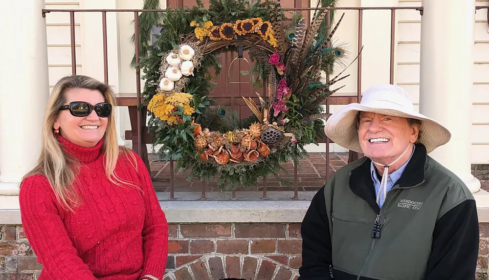 A man and a woman are smiling while standing beside a large ornate wreath adorned with various plants and decorations displayed outside a building with a brick step and railings