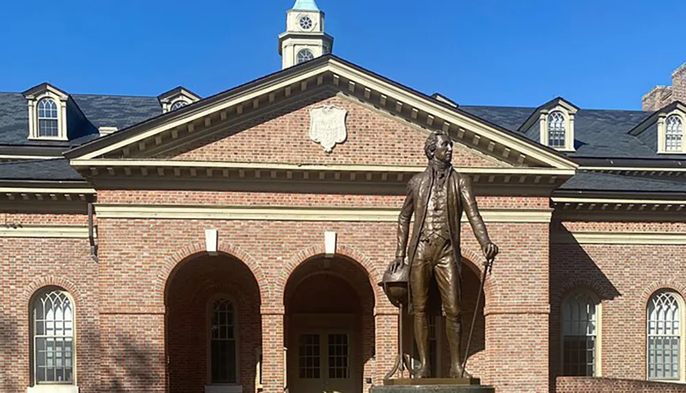 A statue of a historical figure stands in front of a traditional brick building with dormer windows under a clear blue sky