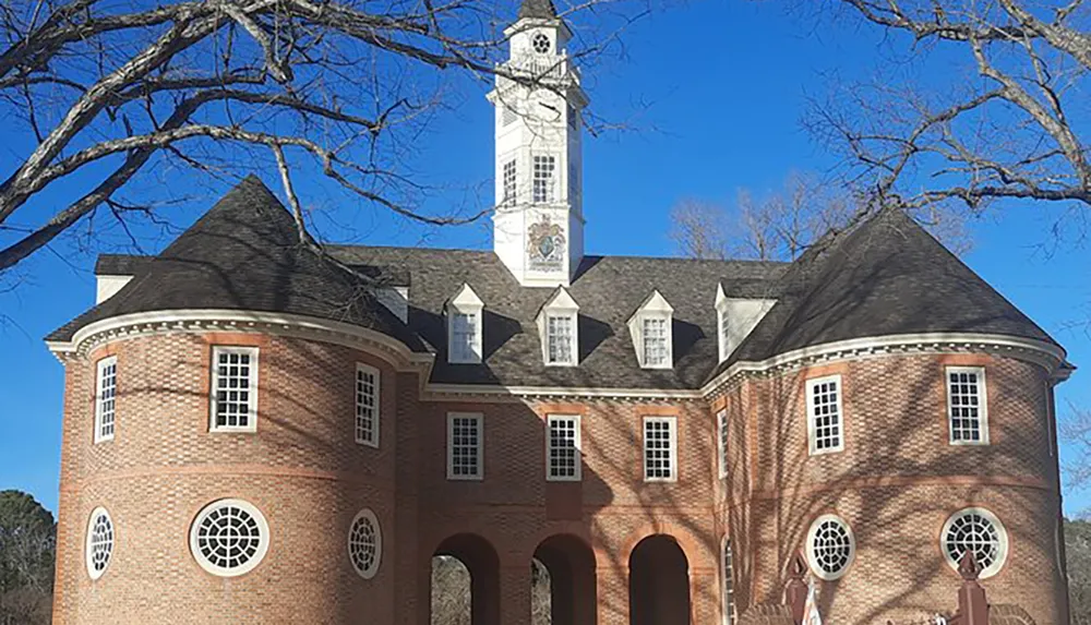 The image shows a colonial-style brick building with a prominent white clock tower adorned by a blue sky and framed by bare tree branches