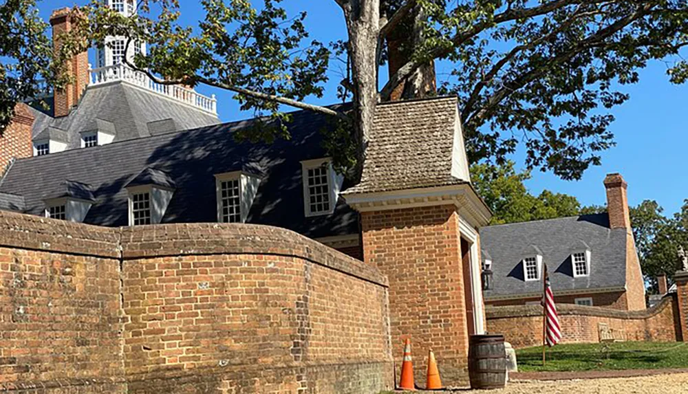 The photo shows a historical colonial-style building with a brick wall in the foreground an American flag and a backdrop of blue sky and trees