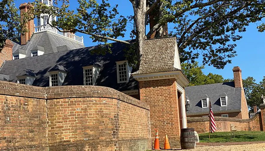 The photo shows a historical colonial-style building with a brick wall in the foreground, an American flag, and a backdrop of blue sky and trees.